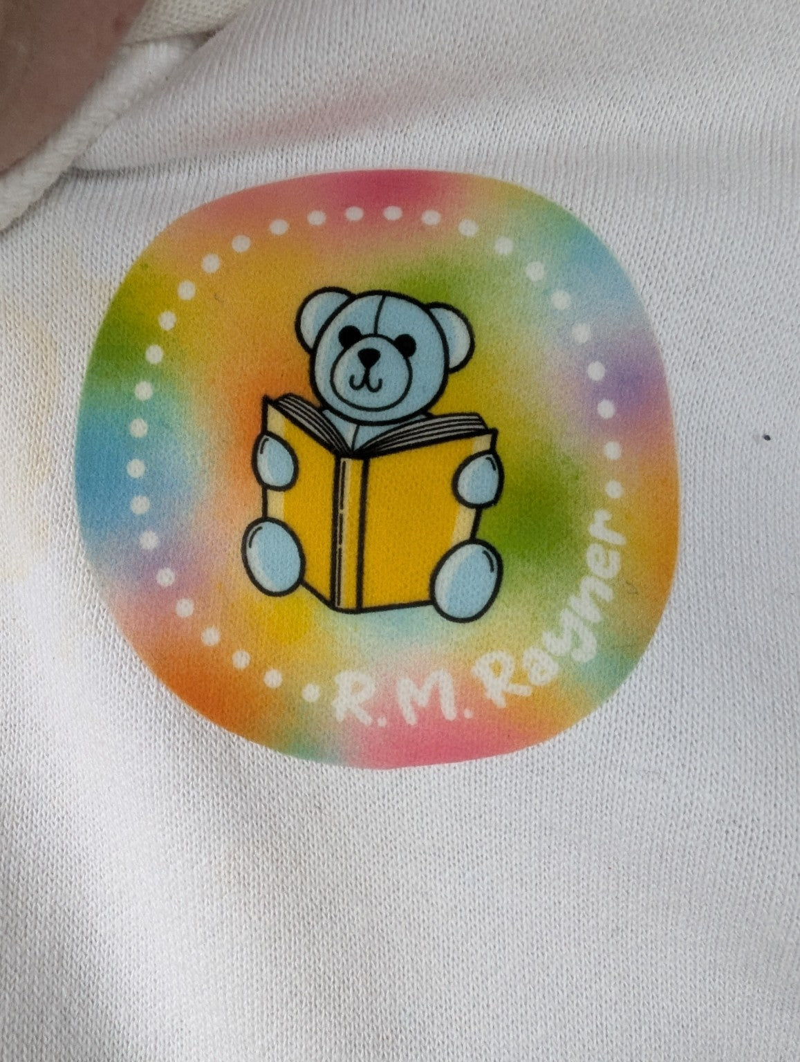 Close-up of the actual pastel rainbow R. M. Rayner teddy reading a yellow book logo printed on a white hoodie