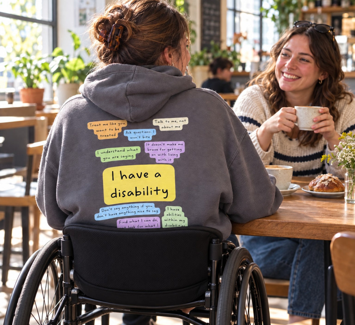 A wheelchair user wearing the I Have a Disability hoodie with its colourful back print, talking with a friend in a café.