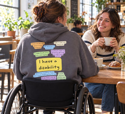 A wheelchair user wearing the I Have a Disability hoodie with its colourful back print, talking with a friend in a café.